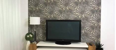 A television on a white stand with a patterned wallpaper, a floor lamp, and a potted plant.