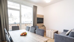 A modern hotel room with a wooden dining table, grey upholstered chairs, a grey sofa, and a small wooden side table. A flat-screen TV is mounted on the wall, and a window offers a view of a snowy landscape. - Apartment for 6 in Fieberbrunn (Fieberbrunn)