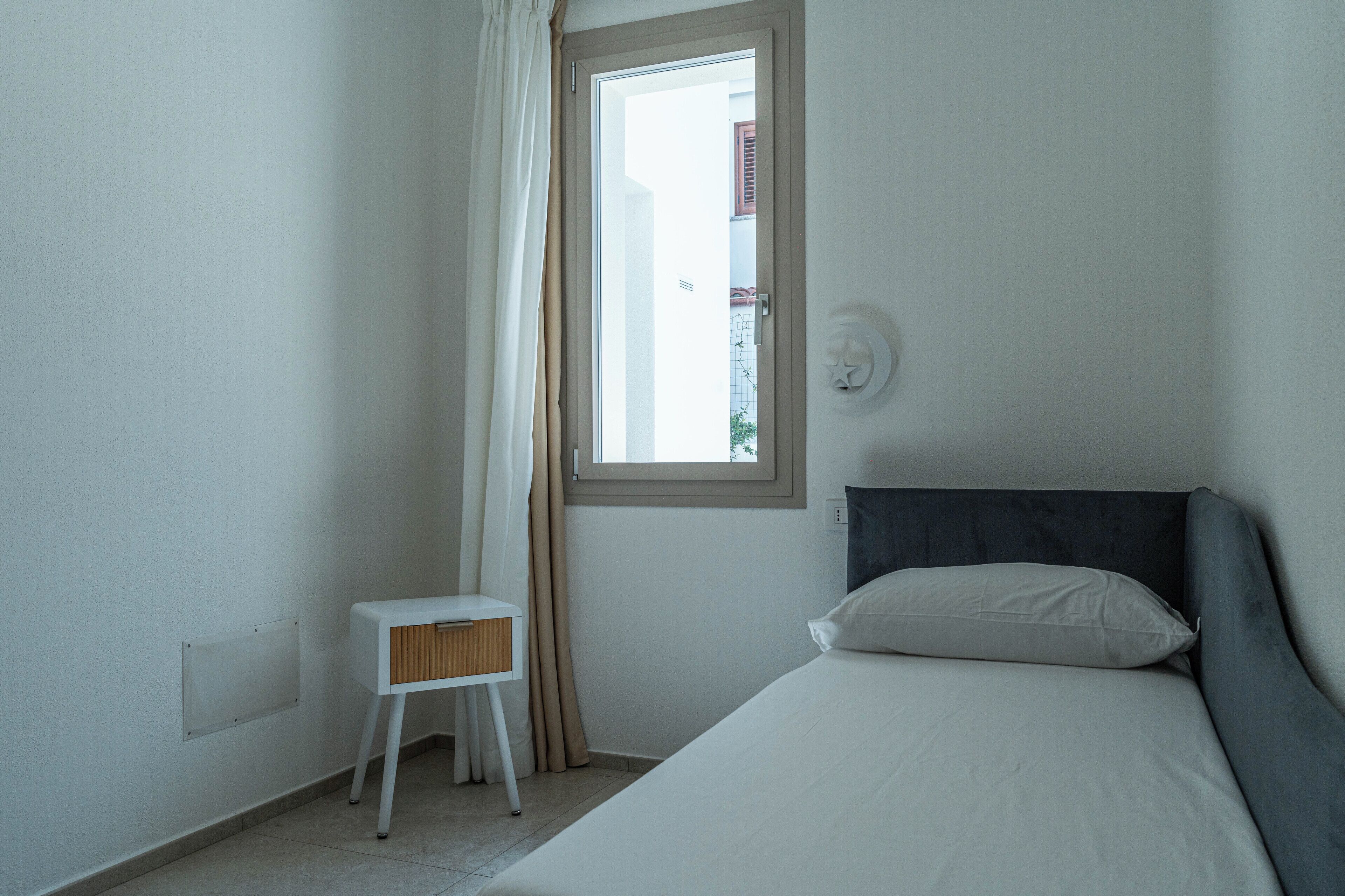 Une petite pièce propre, avec un lit, une petite table, une fenêtre avec des rideaux et un luminaire fixé au mur.