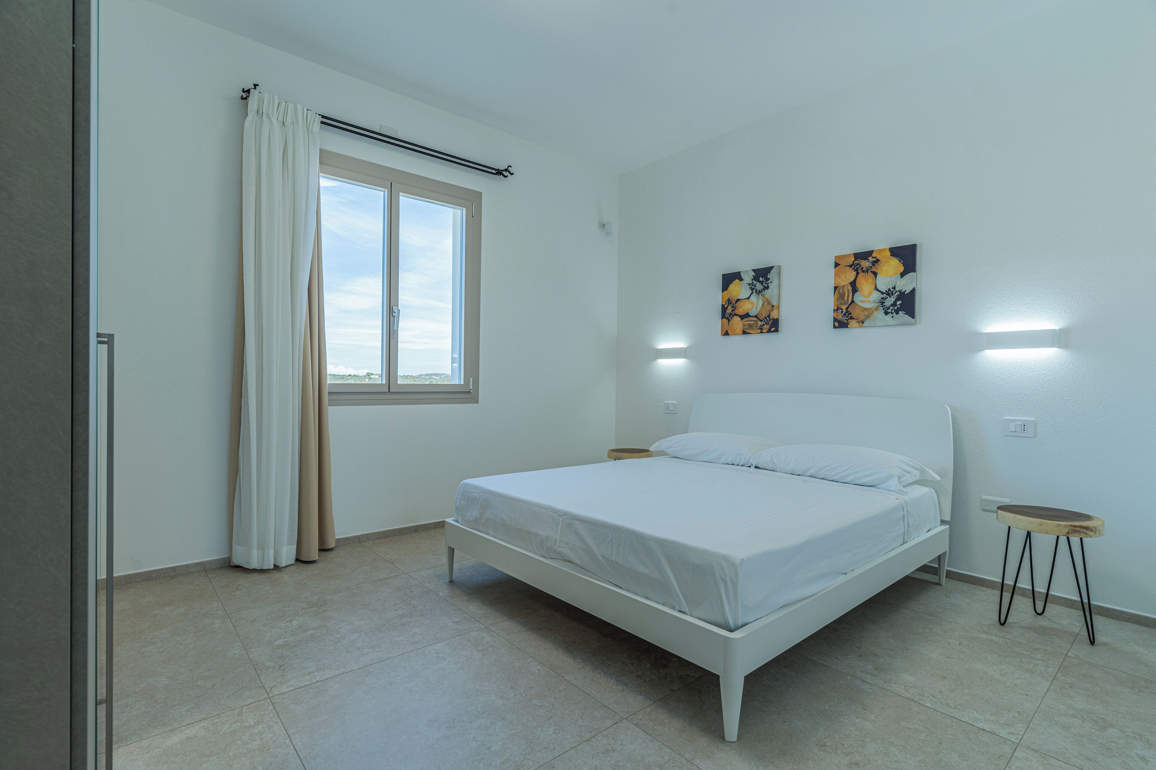 A bedroom with a bed, a window with curtains, a bedside table, and two wall-mounted paintings.