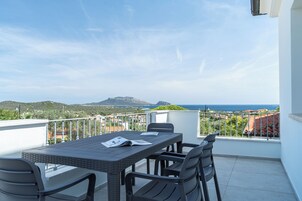 Un balcón con una mesa y sillas, con vista a un paisaje que incluye el mar y las colinas.