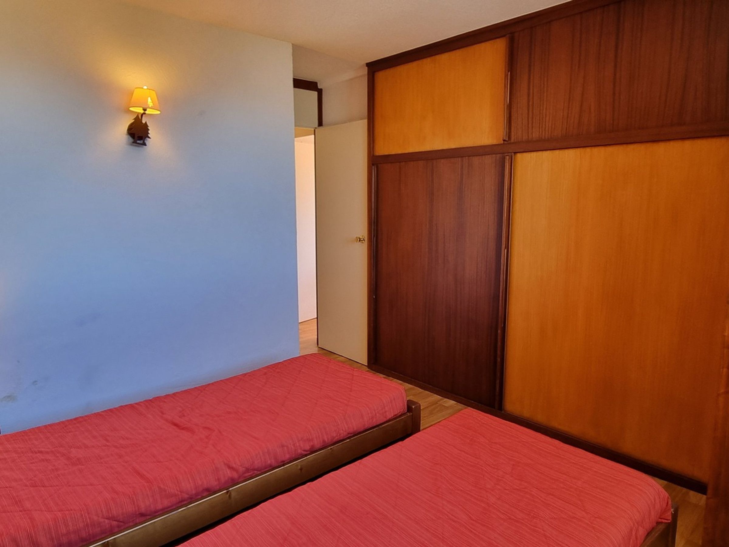 A room with two beds, a wooden wardrobe, and a wall-mounted lamp.