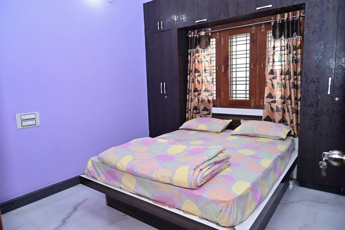 Un dormitorio con cama, paredes moradas, una ventana con cortinas y una puerta con picaporte.
