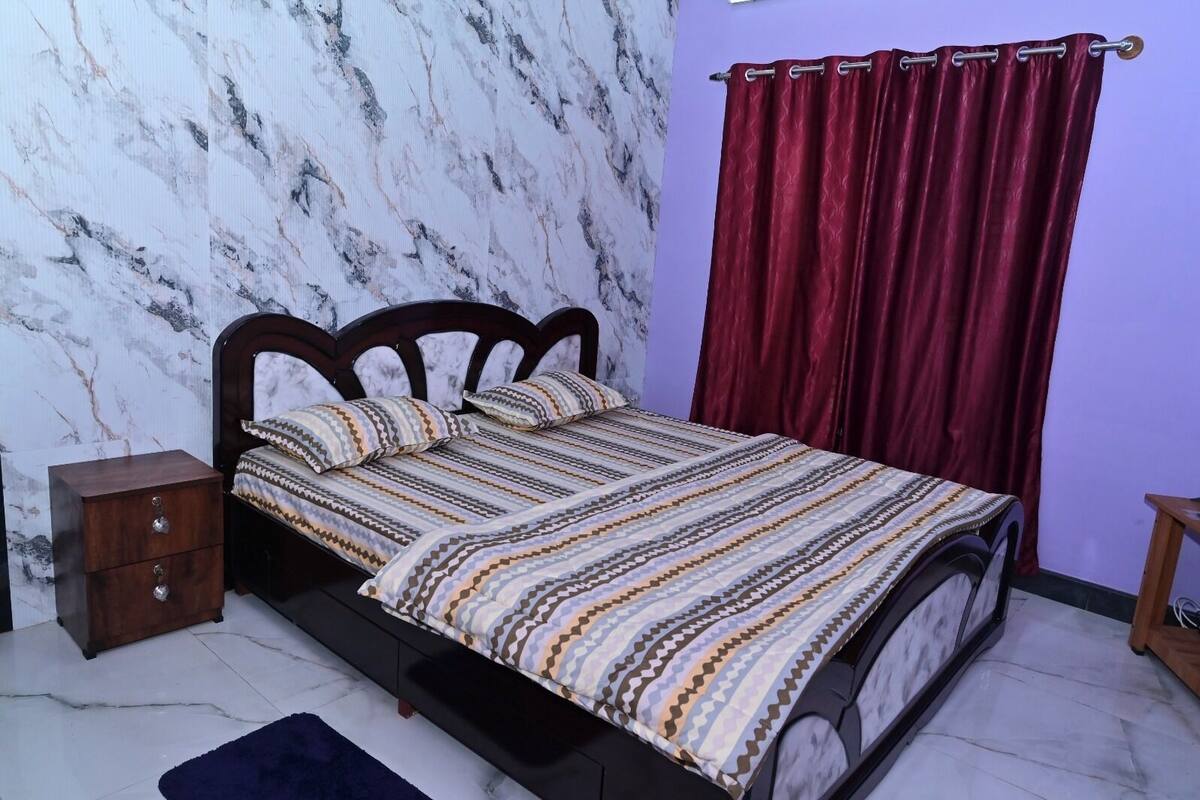 Un dormitorio con cama, mesita de noche, cómoda de madera, pared morada y cortina roja.