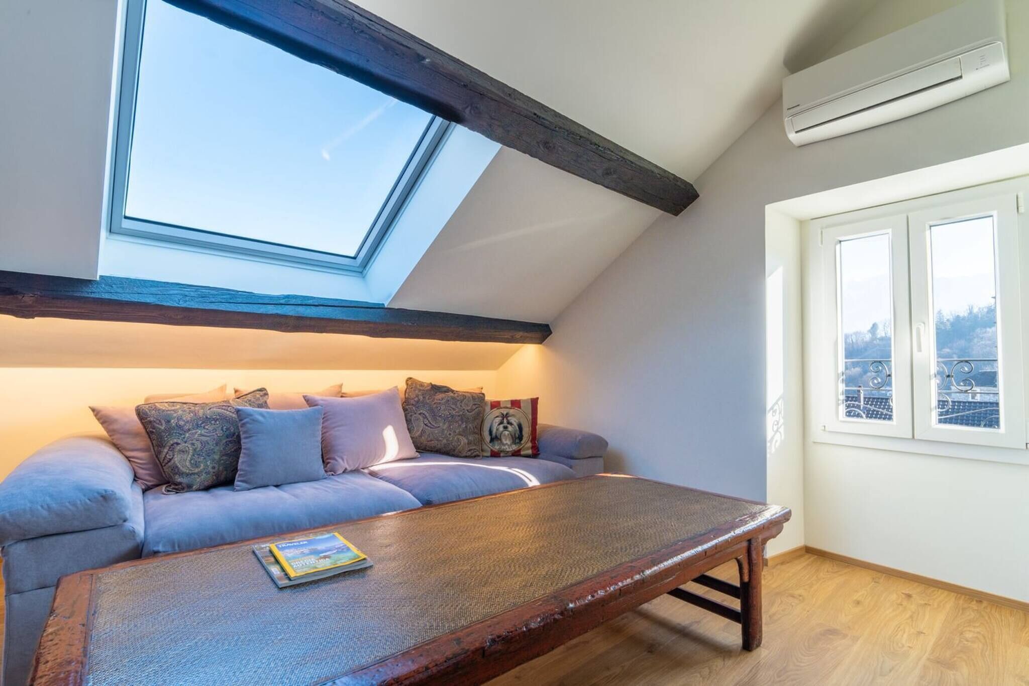 A cozy loft bedroom with a sloped ceiling, a skylight, a wooden coffee table, a sofa with patterned cushions, and a window with a city view.
