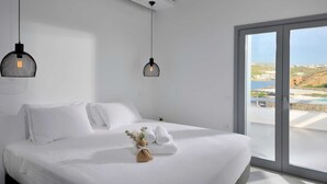 Ein modernes Schlafzimmer mit einem großen Bett, weißen Bettwäsche, zwei hängenden Pendelleuchten und Meerblick durch eine Glastür.