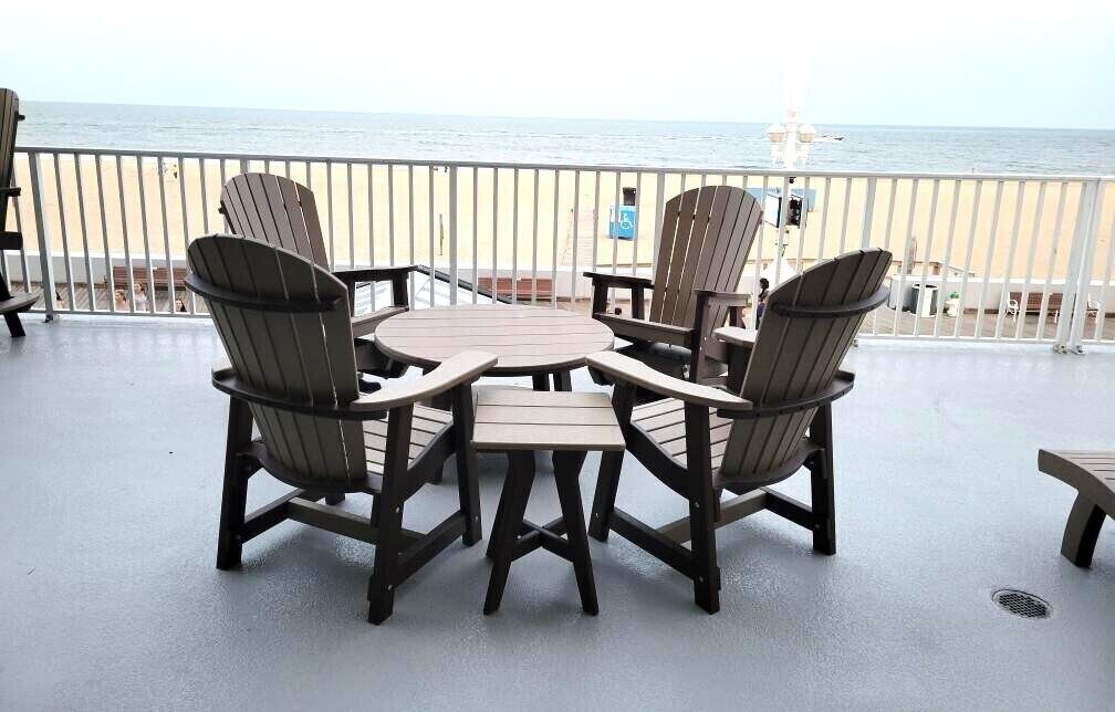 A deck with Adirondack chairs and a round table, overlooking a beach and ocean.