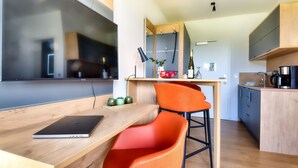 Een moderne keuken met houten kastjes, een eethoek met een laptop op een houten tafel en een feloranje stoel.
