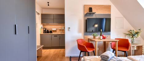 A modern hotel room with a kitchenette, a dining area with two red chairs, a bed with a checkered duvet, and a large TV mounted on the wall.