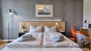 Un lit double avec des oreillers et des serviettes, une table de chevet avec une lampe, un tableau encadré au mur et une chaise avec une couverture.