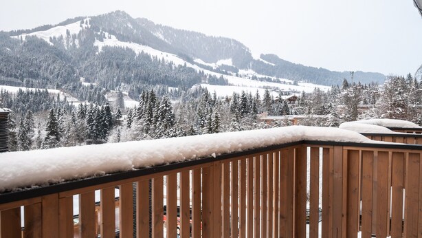 Un balcone in legno con ringhiere imbiancate dalla neve che si affaccia su un paesaggio montano innevato.