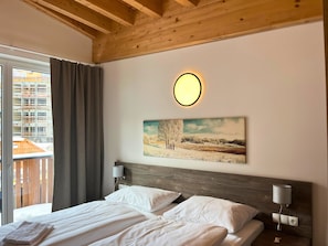 Une chambre d’hôtel avec un plafond en bois, un lit recouvert de draps blancs, un tableau représentant un paysage hivernal et une fenêtre avec des rideaux.