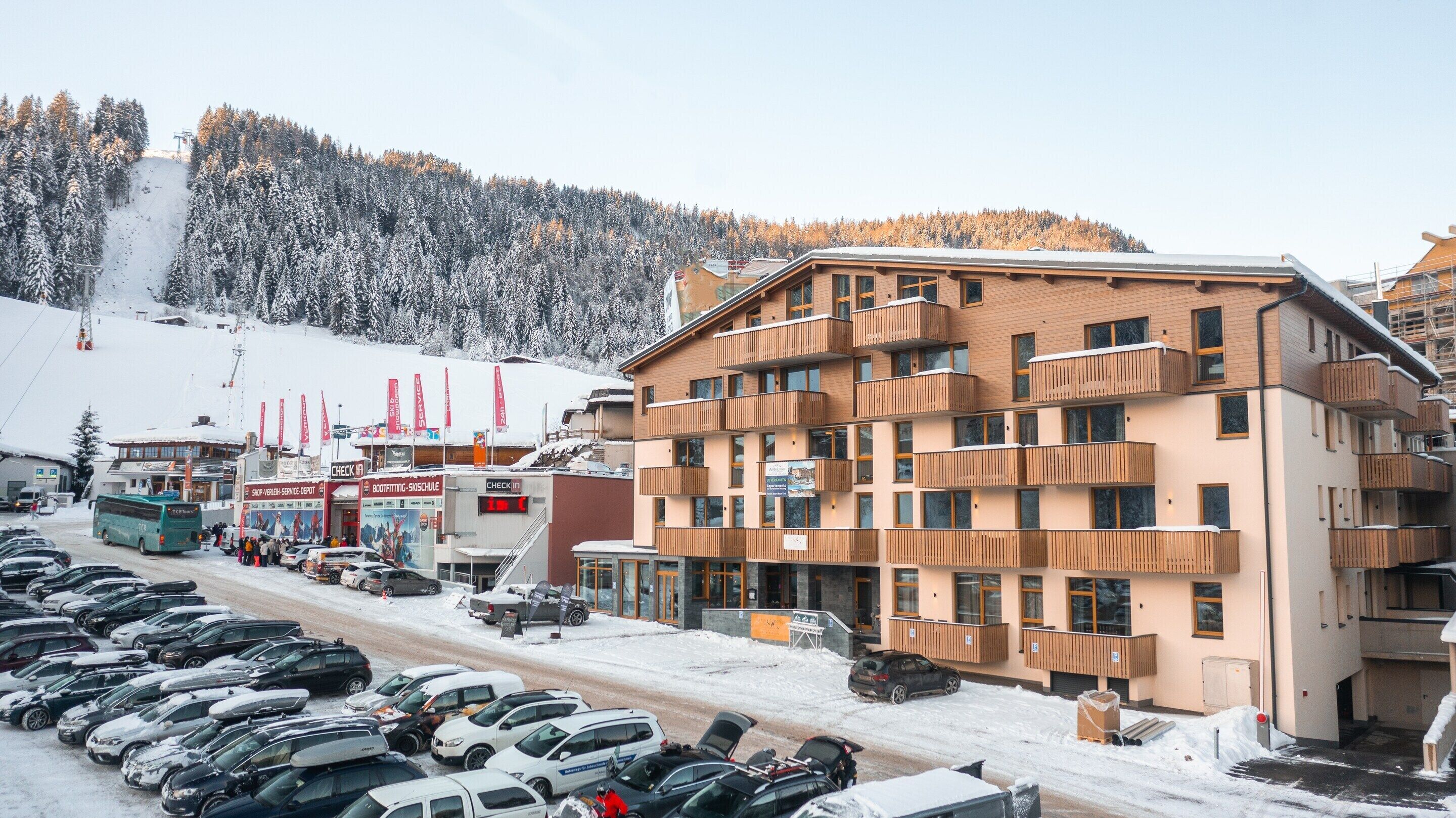 Un hotel de varios pisos con balcones, un telesilla al fondo y un estacionamiento con coches.
