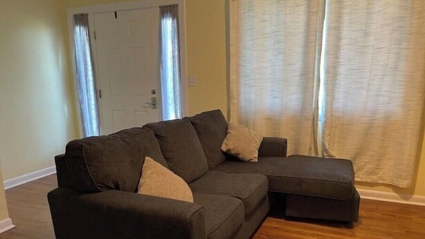 A living room with a dark brown sectional sofa, a small ottoman, and a wooden floor. - Vacation getaway (Morehead City)