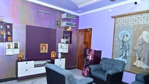 A living room with a purple wall, a wooden door, and a large painting. There are two sofas and a TV stand with decorative items. - Mayur Shines (Anekal)