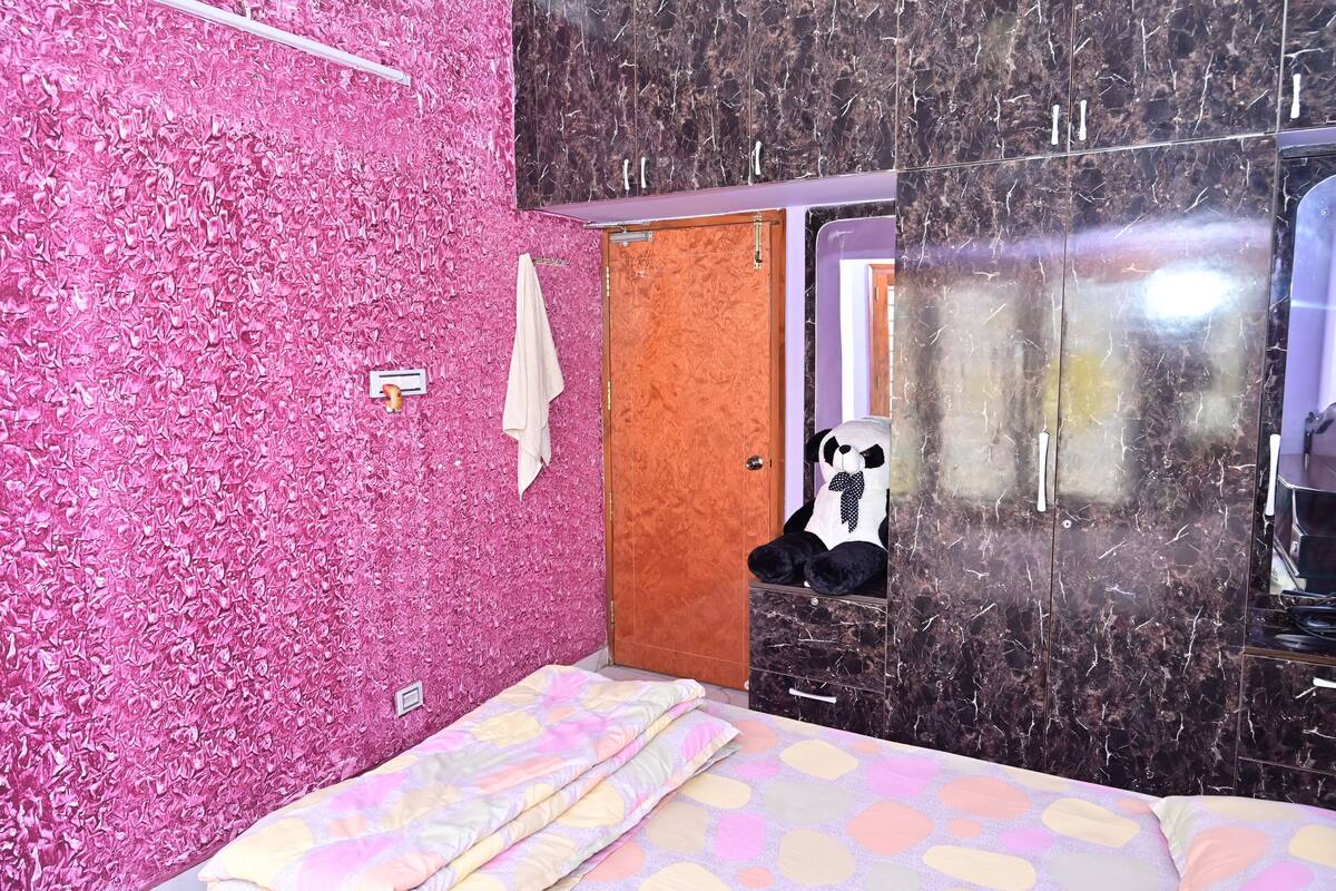 Una habitación con pared rosa con textura, una cama con colcha colorida, un armario con espejo y un peluche de panda sobre el armario.