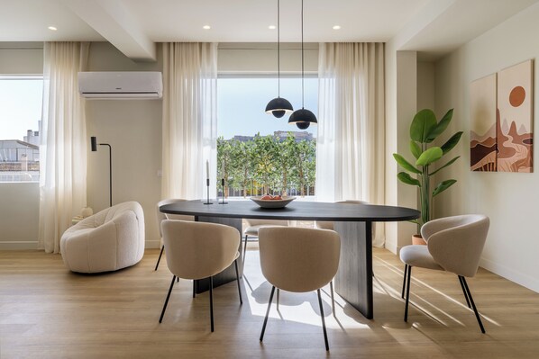 A modern dining area with a round table, two chairs, a potted plant, and a view of greenery outside.