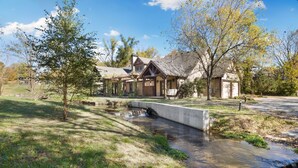 A rustic cabin with a small bridge over a stream, surrounded by trees and a clear sky. - Whispering Brook-Outdoor oasis w/stream-Sleeps 16 (Springdale)
