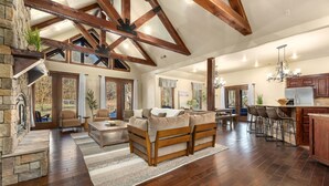 A spacious living room with a fireplace, wooden beams, a stone accent wall, and a large window overlooking a deck. - Whispering Brook-Outdoor oasis w/stream-Sleeps 16 (Springdale)