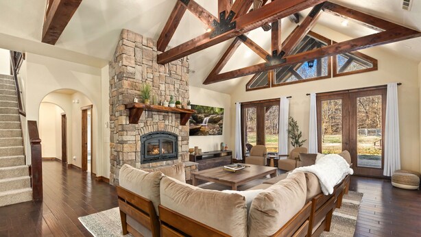 A spacious living area with a stone fireplace, wooden beams, a large sofa, and a view of the outdoors through sliding glass doors. - Whispering Brook-Outdoor oasis w/stream-Sleeps 16 (Springdale)