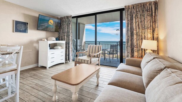 A hotel room with a balcony, a sofa, a dining table, and a TV. - Ocean Forest Plaza 708 - Oceanfront (Myrtle Beach)