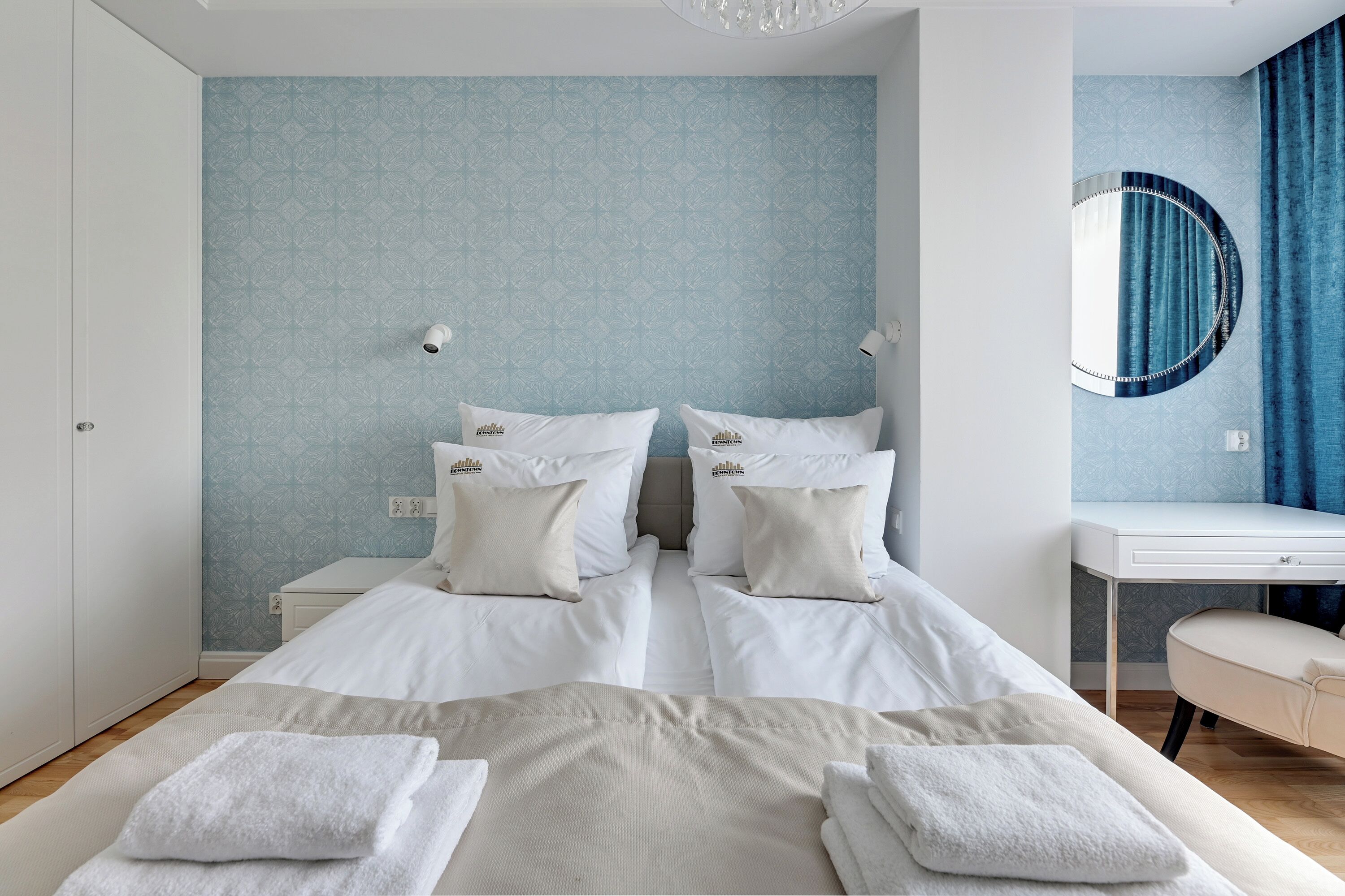 A double bed with white linens and two pillows, a bedside table, a desk with a chair, and a round mirror on the wall.