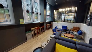 A modern lounge area with a mix of seating options including sofas and chairs, a coffee table, and a small dining table. Large windows provide natural light and views of the street. - Student Only Shoreditch (London)