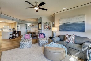 A living room with a sofa, ottomans, a coffee table, a dining area with chairs, a kitchen in the background, and a ceiling fan.