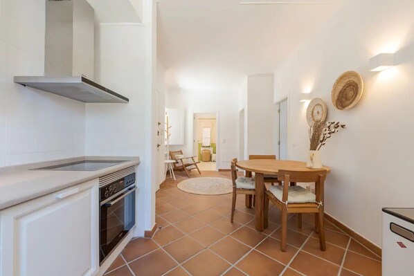 A modern kitchen with white cabinets, a built-in oven, and a stainless steel range hood. There is a dining area with a wooden table and chairs, and a small rug on the floor.