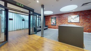 A lobby with a wooden floor, a reception desk, a glass door entrance, and a brick wall with a poster. - Student Only Will Wyatt Court (London)