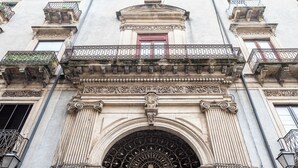 A building facade with ornate architectural details, including a central arched entrance with decorative metalwork and multiple balconies with iron railings. - Palazzo Bicocca Rooms (Catania)