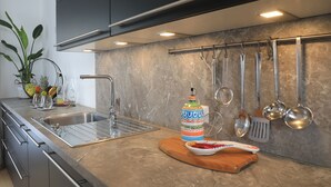 Une cuisine moderne avec un évier en acier inoxydable, un panier de fruits, une bouteille colorée et divers ustensiles de cuisine accrochés au mur.