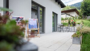 Un bâtiment moderne doté d’un patio comprenant une table et des chaises, un petit jardin avec des plantes et deux affiches présentées sur des chevalets.