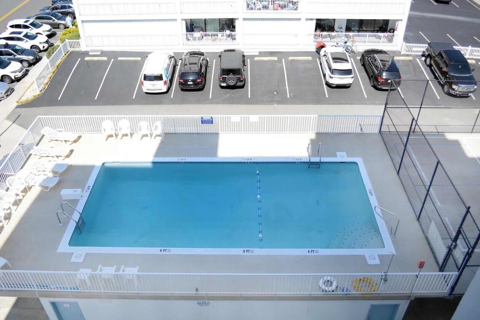 Eine Vogelperspektive auf ein Schwimmbecken mit angrenzenden Parkplätzen und einem Gebäude mit weißer Fassade.