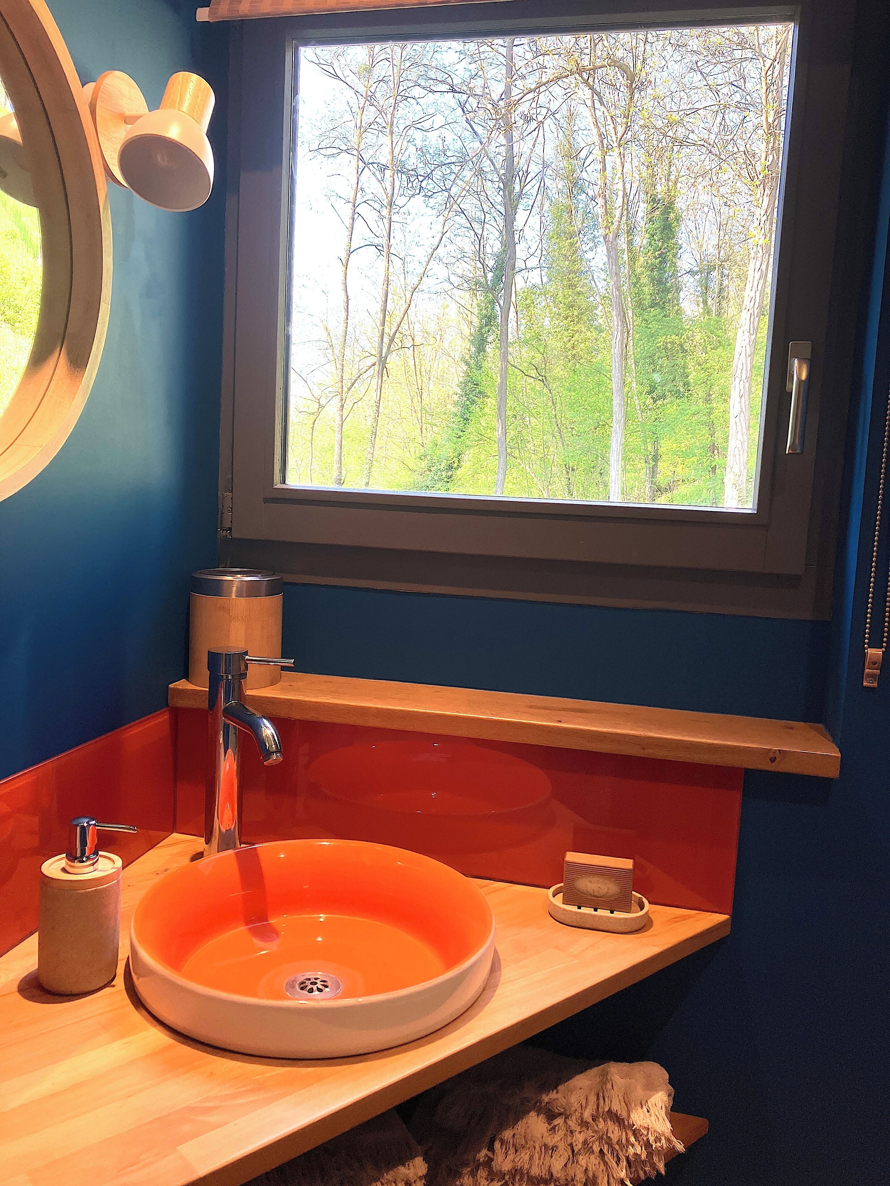 Une salle de bain avec un meuble-lavabo en bois, un lavabo orange, un miroir et une fenêtre donnant sur des arbres.