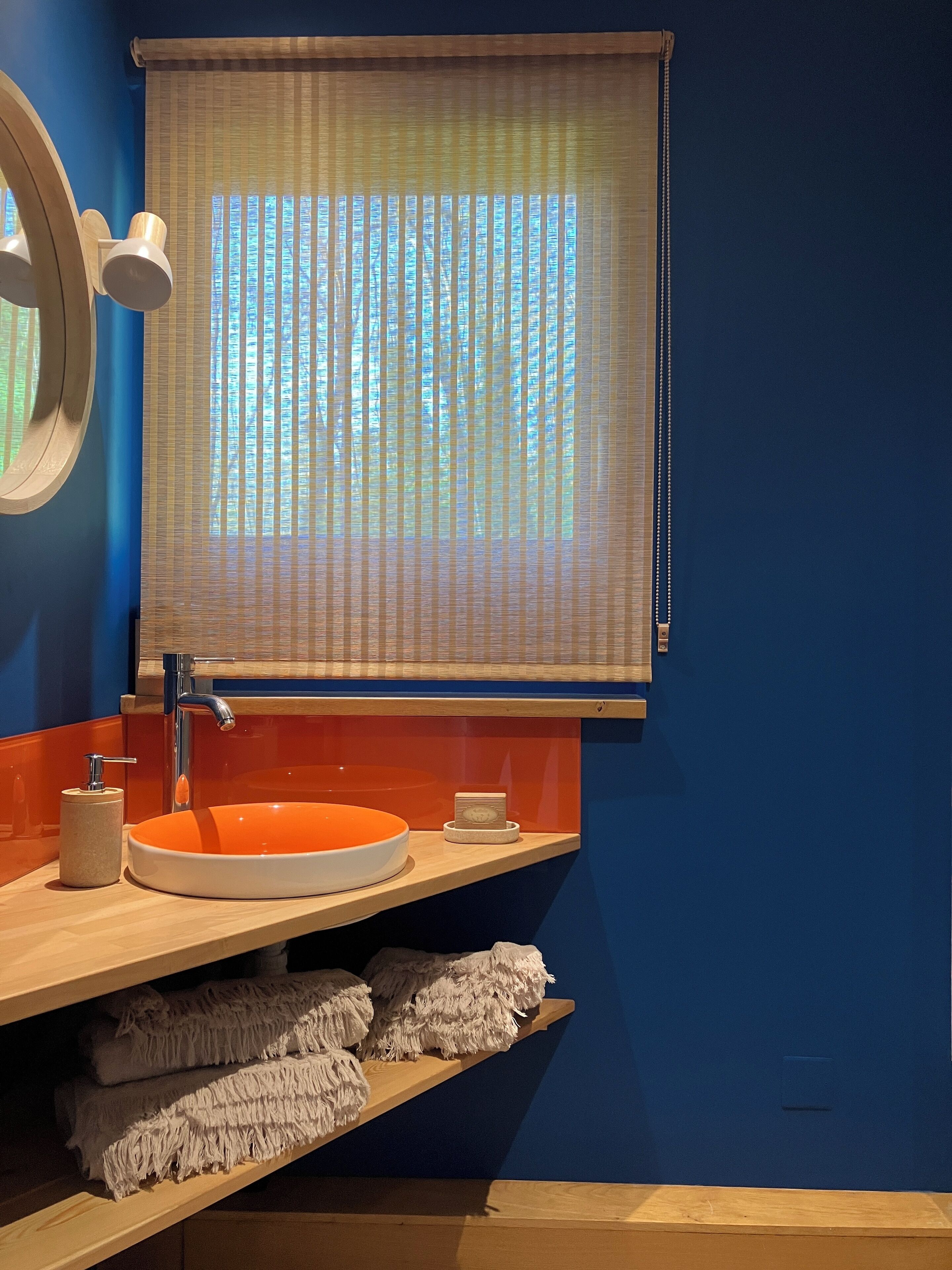 Een badkamer met een ronde spiegel, een wastafel met een oranje wasbak, een houten plank met handdoeken, en een raam met zonwering.