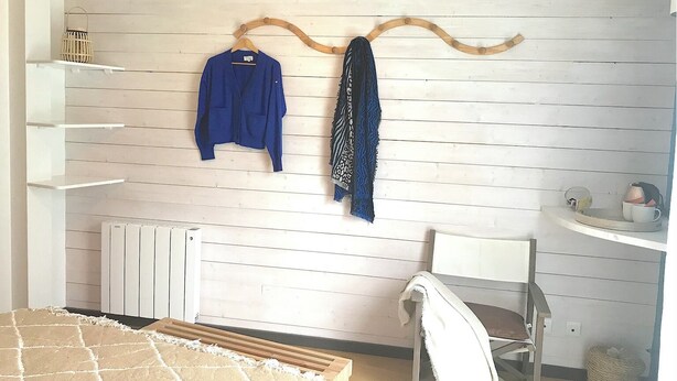 A room with a white wooden wall, a white radiator, a white chair, a white shelf, and a hanging wooden rack with clothes. - A bed and breakfast in the woods (Pommiers)