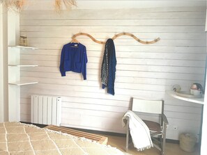 Una habitación con pared de madera blanca, un radiador blanco, una silla blanca, una estantería blanca y un perchero de madera con ropa colgada.