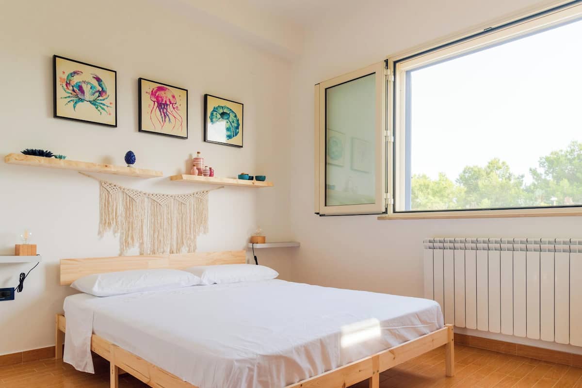 A bedroom with a wooden bed, white bedding, a large window, and wall-mounted shelves with decorative items.