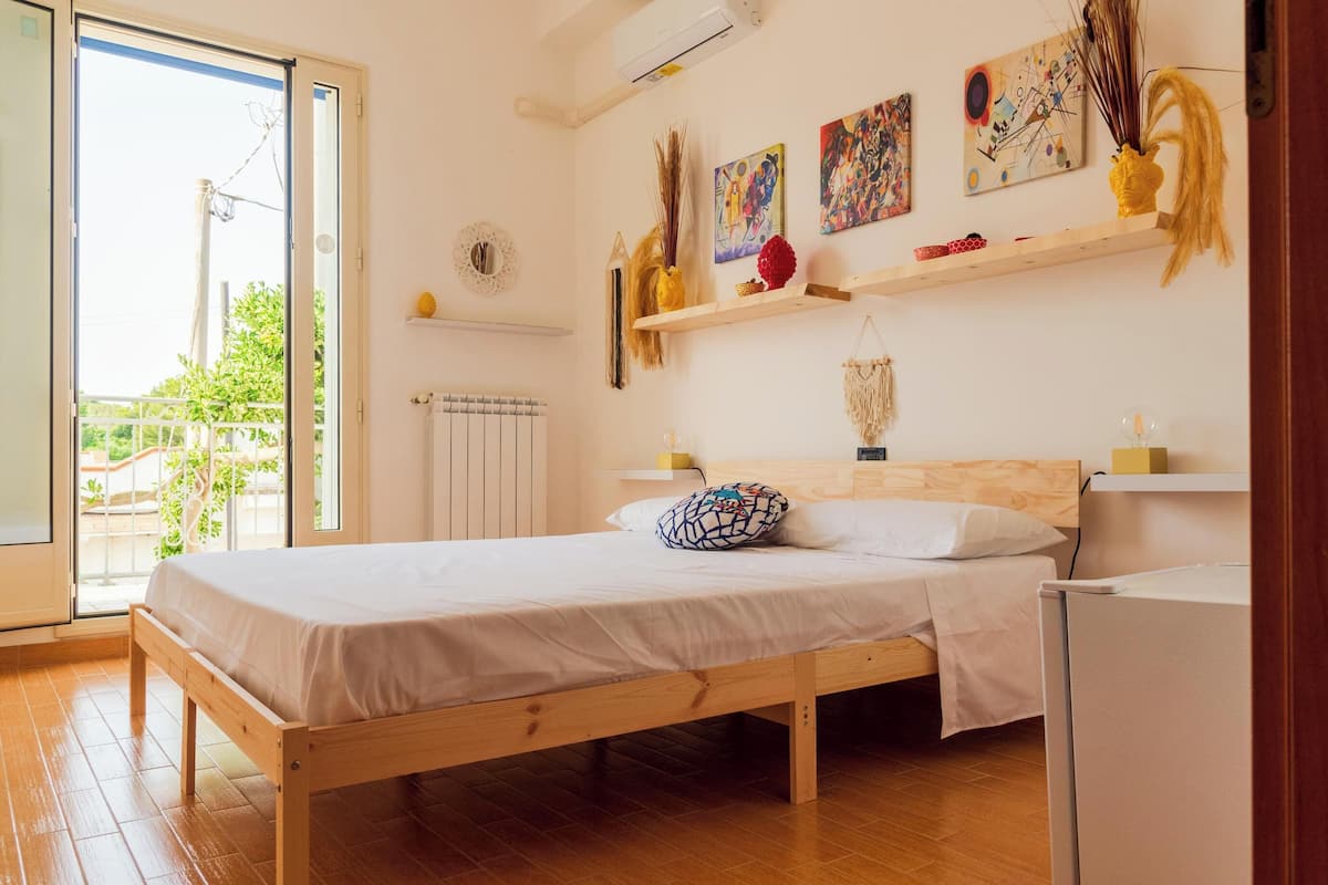 A bedroom with a wooden bed, white bedding, a small bedside table, a wall-mounted shelf with decorative items, and a window with a view of greenery.