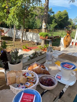 An outdoor breakfast setting with croissants, yogurt, and grapes on a table, surrounded by potted plants and a scenic garden view.
