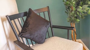 Una silla con cojín y almohada oscura, reposabrazos de madera y una planta al fondo.