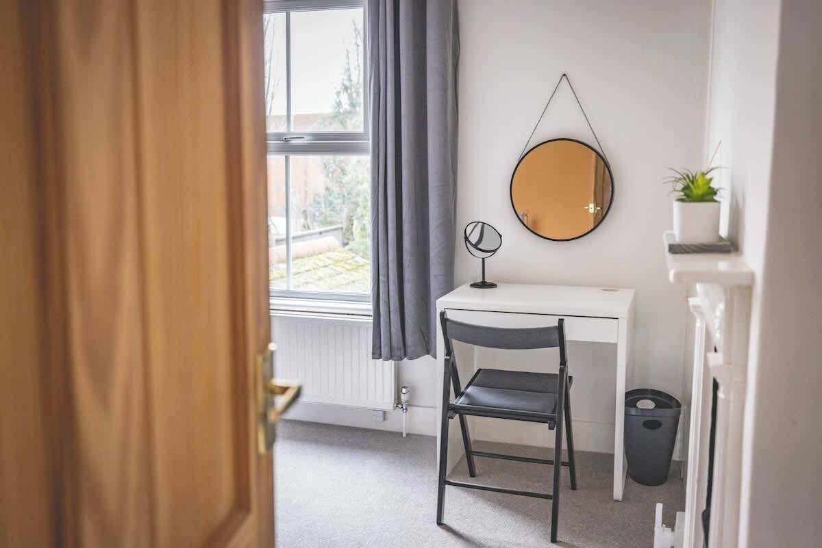 A room with a white desk, a chair, a round mirror, a potted plant, and a window with curtains.