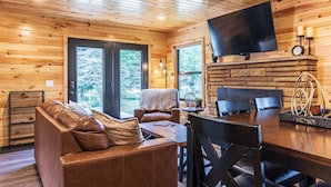 A cozy cabin interior with a wooden ceiling, a flat-screen TV mounted above a stone fireplace, a leather sofa, and a dining table with chairs. - Fall on Whitefish Bay | Private Beach, Fireplace (Paradise)