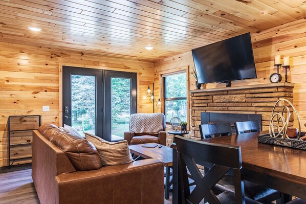 A cozy cabin interior with a wooden ceiling, a flat-screen TV mounted above a stone fireplace, a leather sofa, and a dining table with chairs.