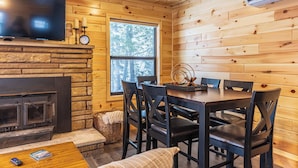 A cozy cabin interior with a stone fireplace, wooden walls, a dining table with chairs, a television, and a window with a view of trees. - Fall on Whitefish Bay | Private Beach, Fireplace (Paradise)