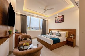 Quarto de hotel moderno com uma cama grande, uma escrivaninha, uma cadeira, um ventilador de teto e uma janela com vista para a cidade.