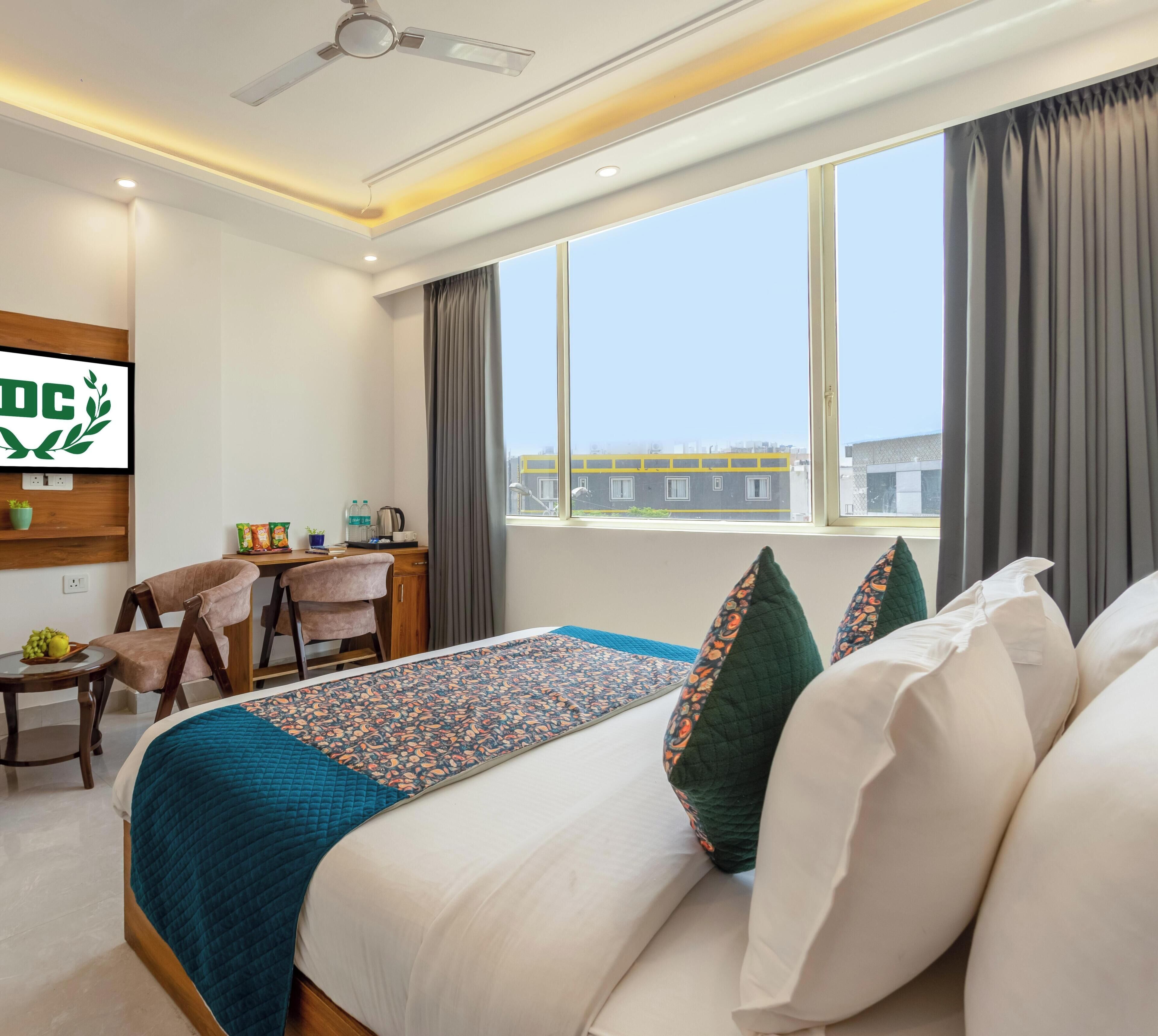 A hotel room with a bed, a desk, a chair, a TV, and a window with a view of buildings.