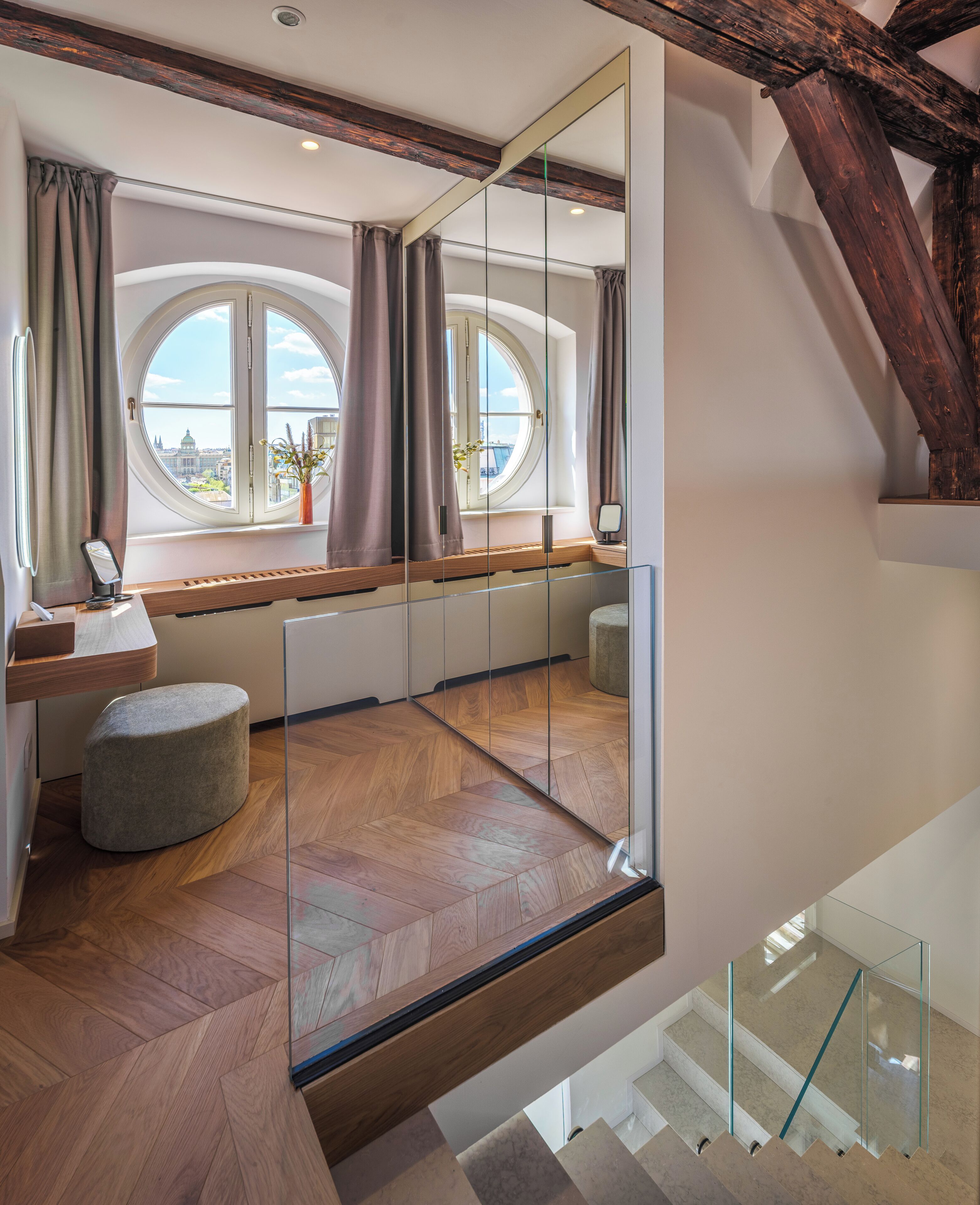 A modern room with a large window, a wooden bench, a round ottoman, and a glass railing.
