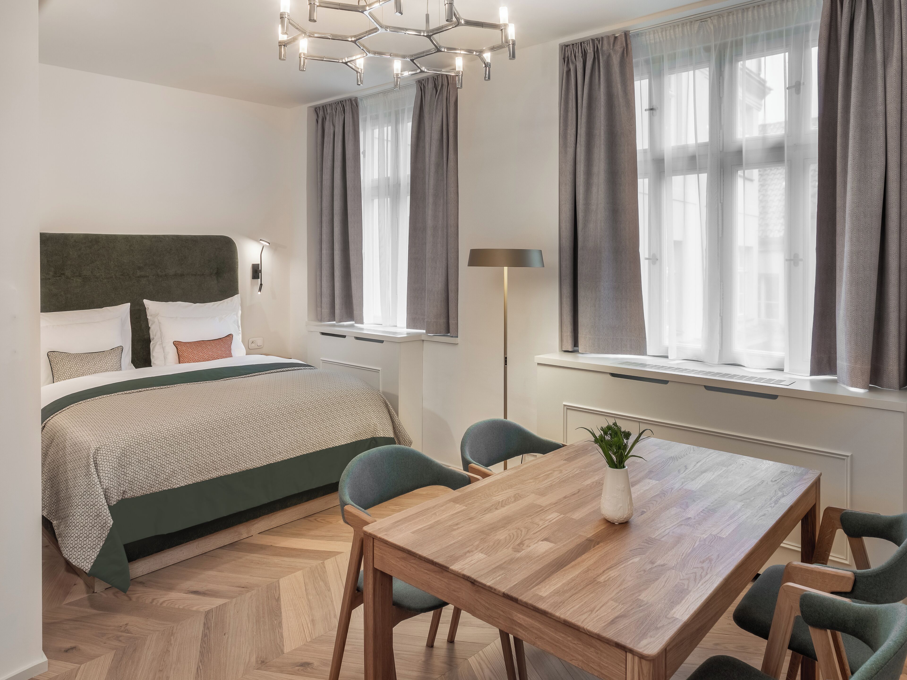 Moderni hotellihuone, jossa on suuri sänky, ruokapöytä ja tuoli. Huoneessa on ikkuna, jossa on verhot, lattiavalaisin ja kattovalaisin.